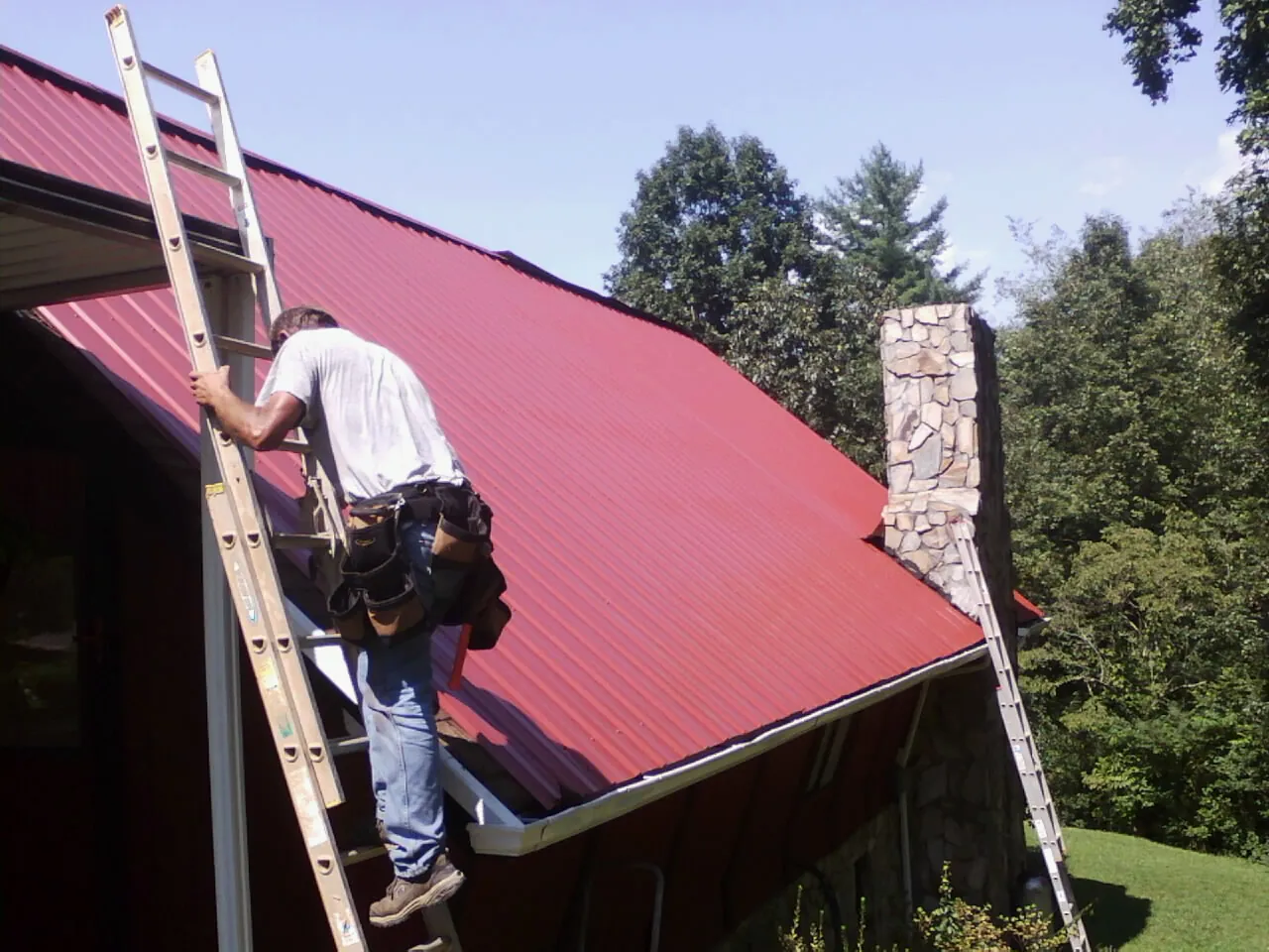 Roofer on a ladder beside a red metal roof and stone chimney — Soffit & Fascia Repair by Roofing Brothers in Newell